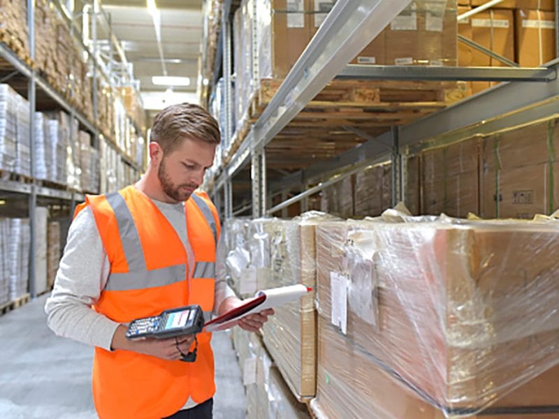 Man in factory hall wearing safety vest holding clipboard and barcode scanner